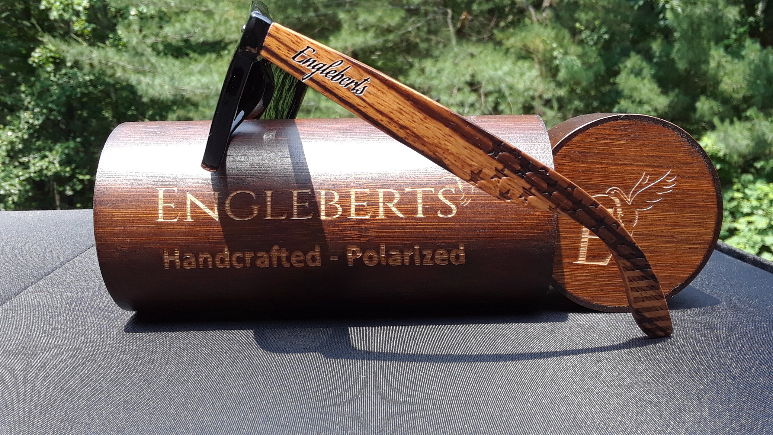 Wooden sunglasses with "Engleberts" on arm rest on a brown engraved case against greenery.