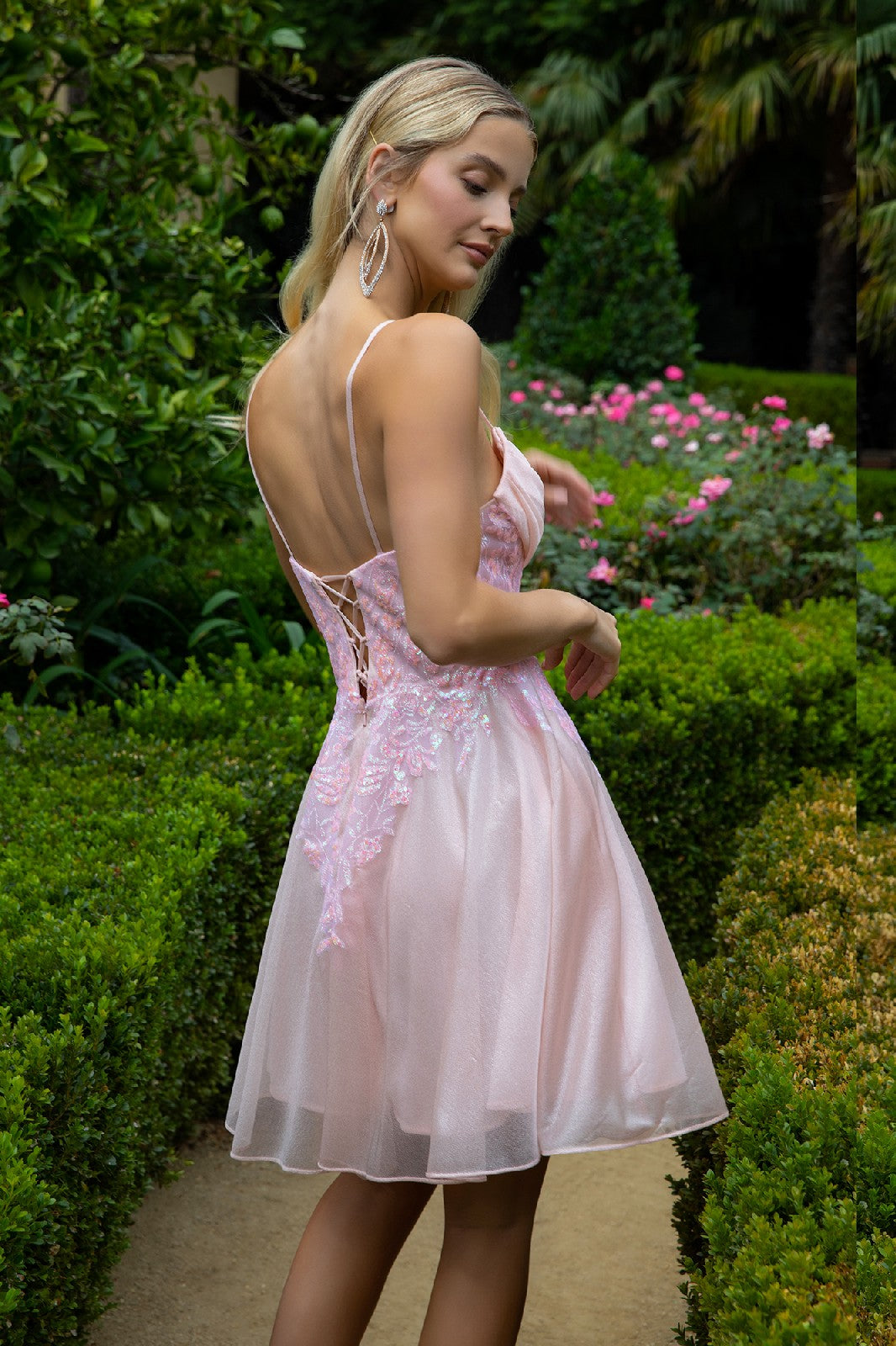 Woman in a pink floral dress in a garden, surrounded by green shrubs and pink flowers.