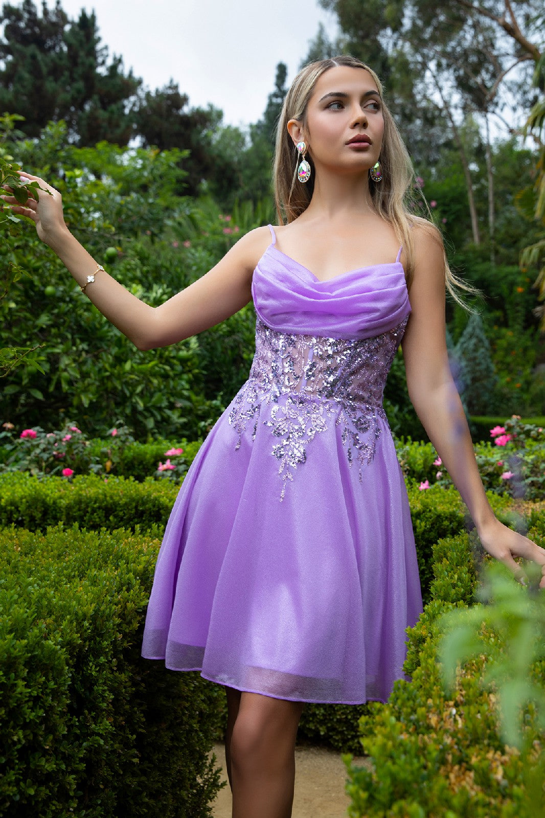 Woman in purple floral dress standing in garden with green hedges and lush foliage.