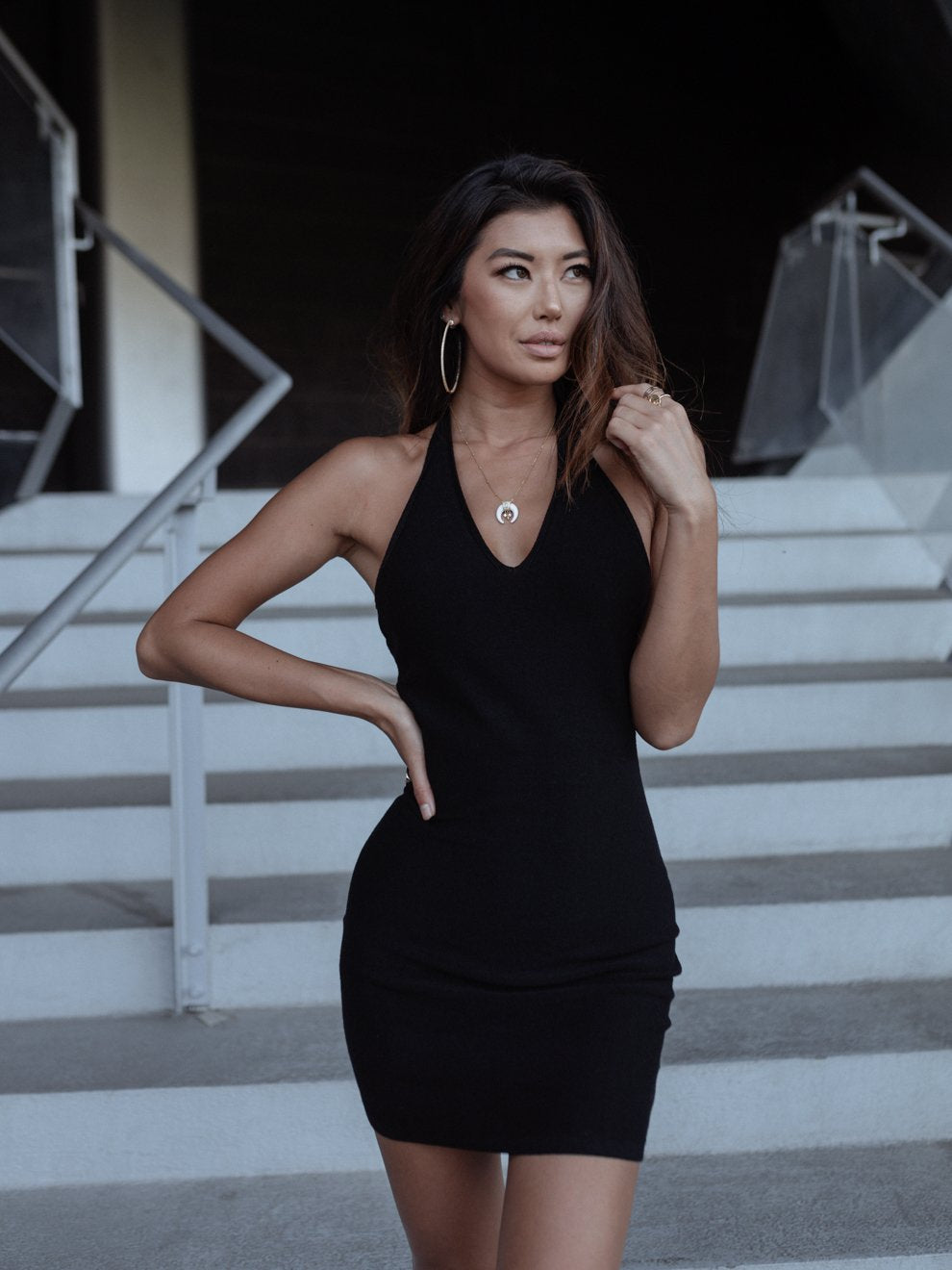Woman in a black dress with large hoop earrings standing on stairs, hand on hip, looking sideways.
