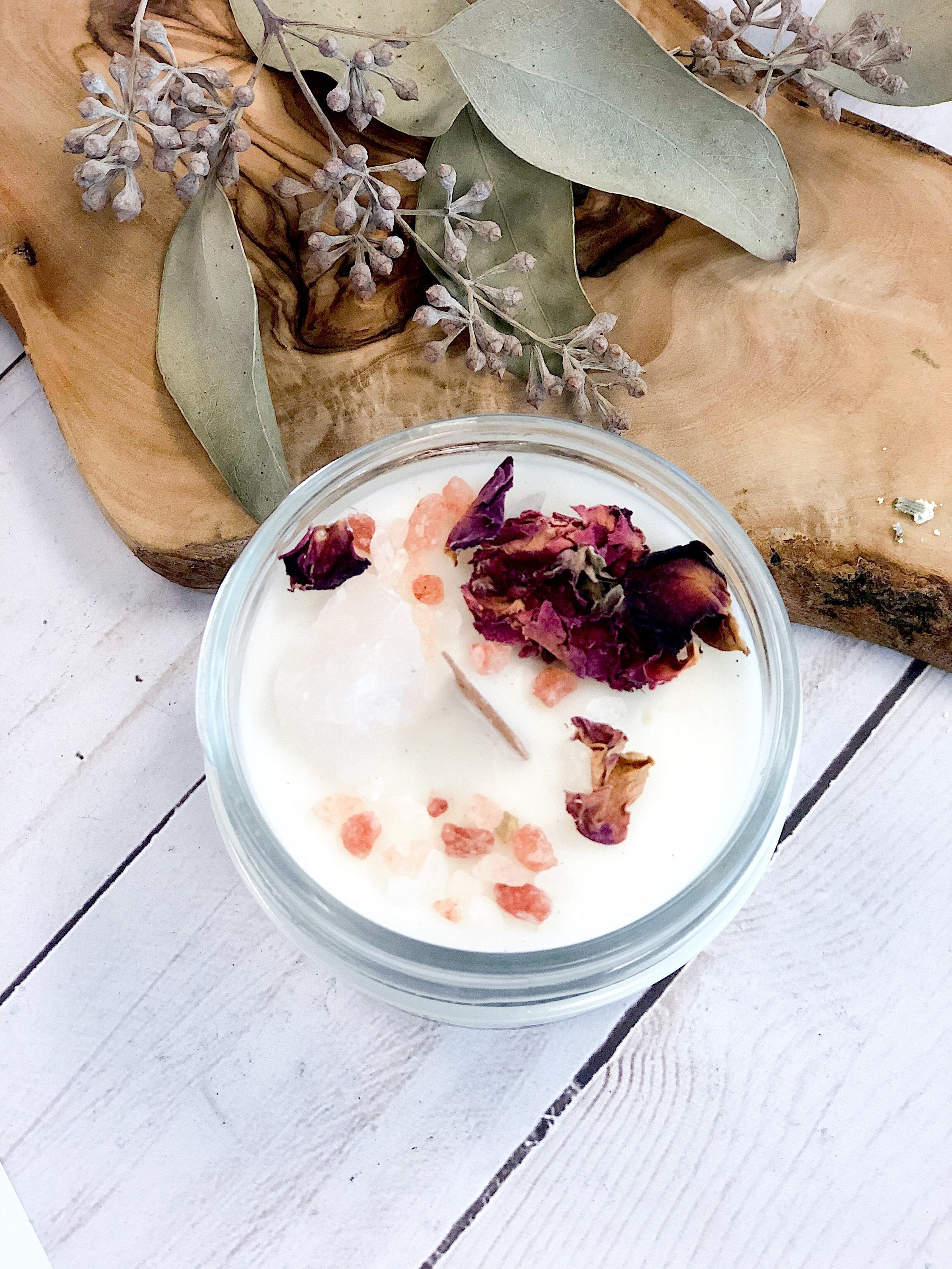 A glass jar with white cream topped with pink salt and dried rose petals on wood with leaves.