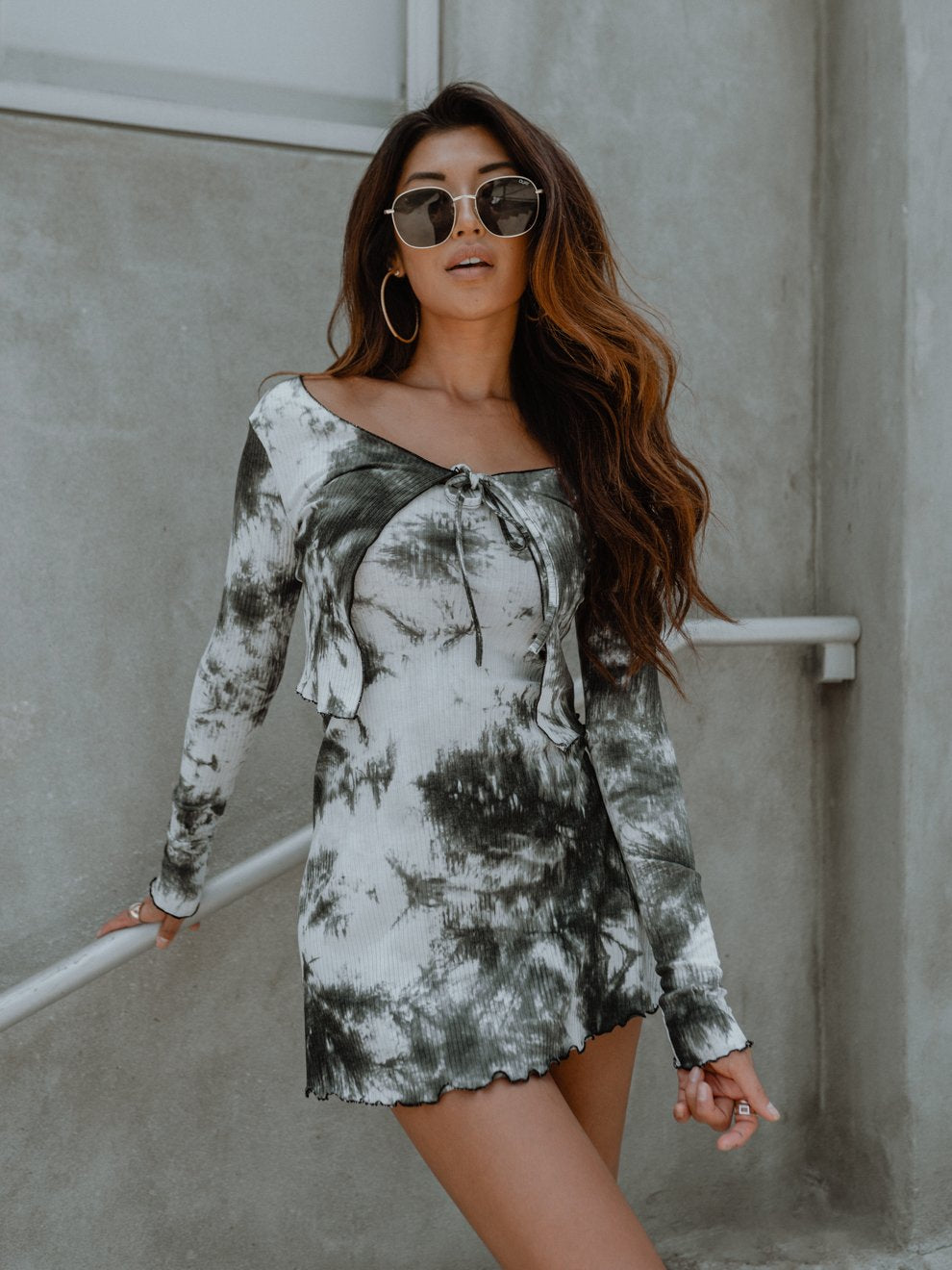 Woman in sunglasses wearing a black and white tie-dye dress stands against a concrete wall.