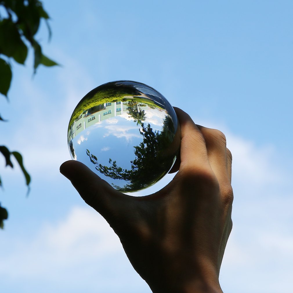 Hand holding a glass ball reflecting a house, sky, and trees; against a bright blue sky backdrop.