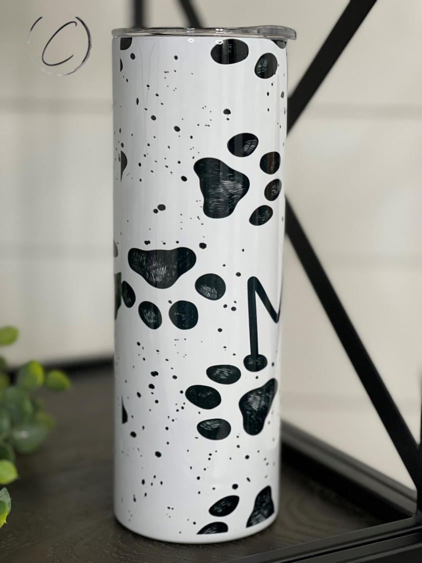 White tumbler with black paw prints and splatters, on a dark surface with greenery in the background.