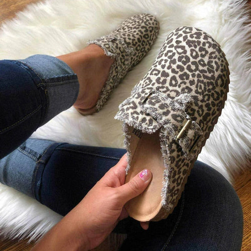 Leopard print clogs with frayed edges held by a person in jeans, resting on a white fluffy rug.