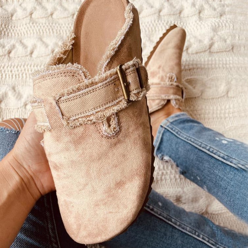 Beige slip-on shoe with frayed edges and brass buckle, held by hand over a knit background.