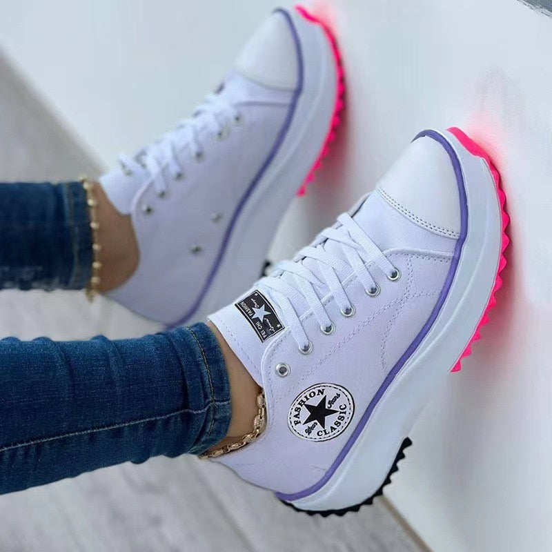 White sneakers with purple trim and neon pink soles, worn with blue jeans against a white wall.