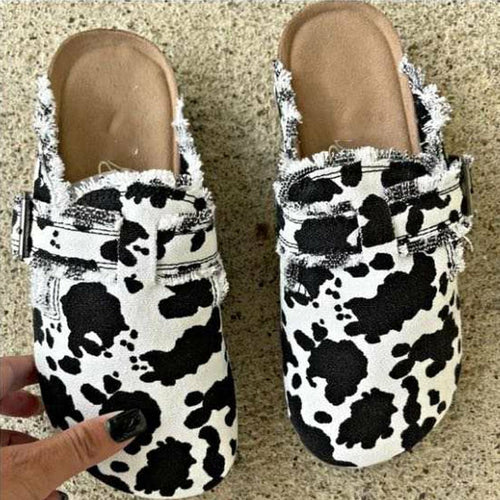 Black and white cow-print slip-on shoes with frayed edges on a textured beige surface.