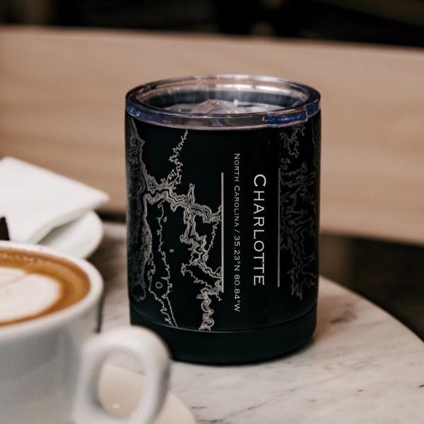 Black tumbler with map design and "Charlotte" text, on marble table near a cappuccino.