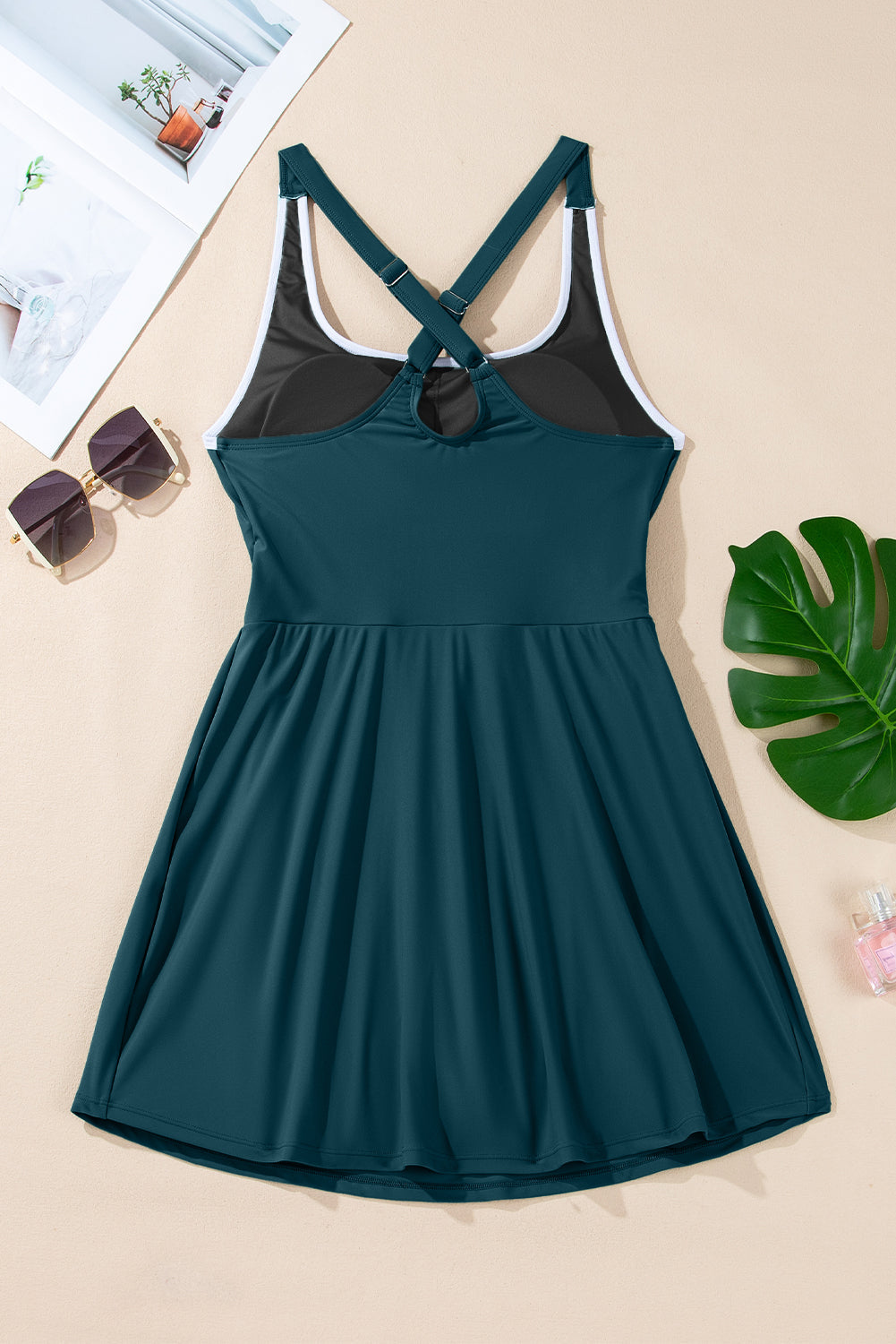 Teal dress with crisscross straps on beige background, near sunglasses, magazine, and a leaf.