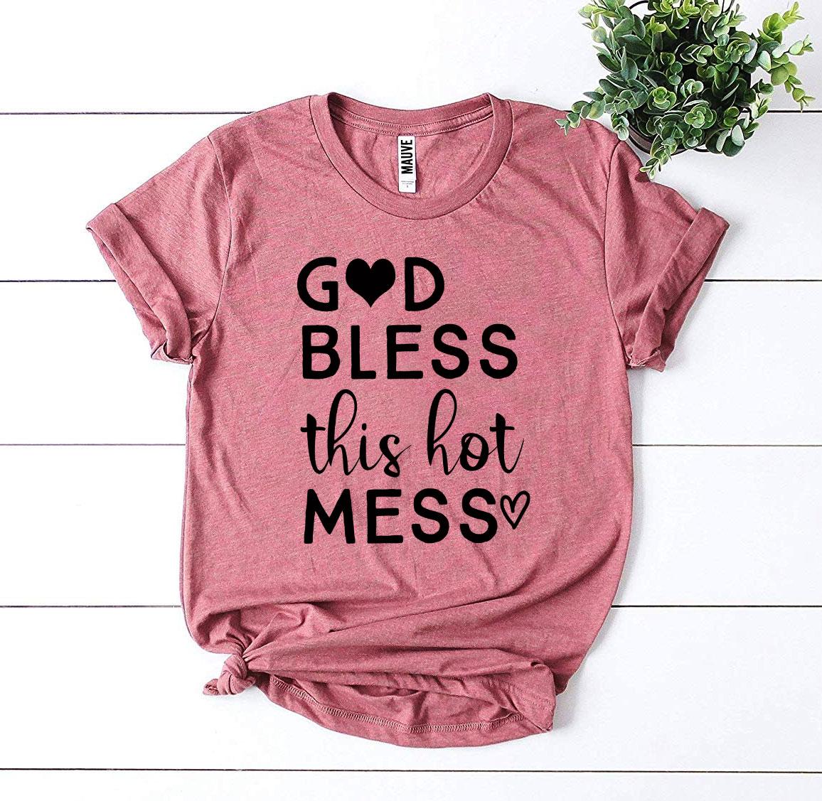 Pink T-shirt with "God Bless This Hot Mess" text and heart icon, placed on white wooden surface.
