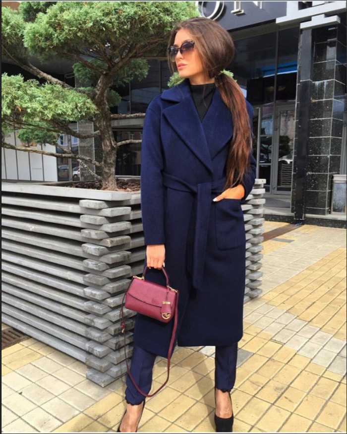 Woman in a navy coat and pants, holding a burgundy bag, stands by a tree and building.