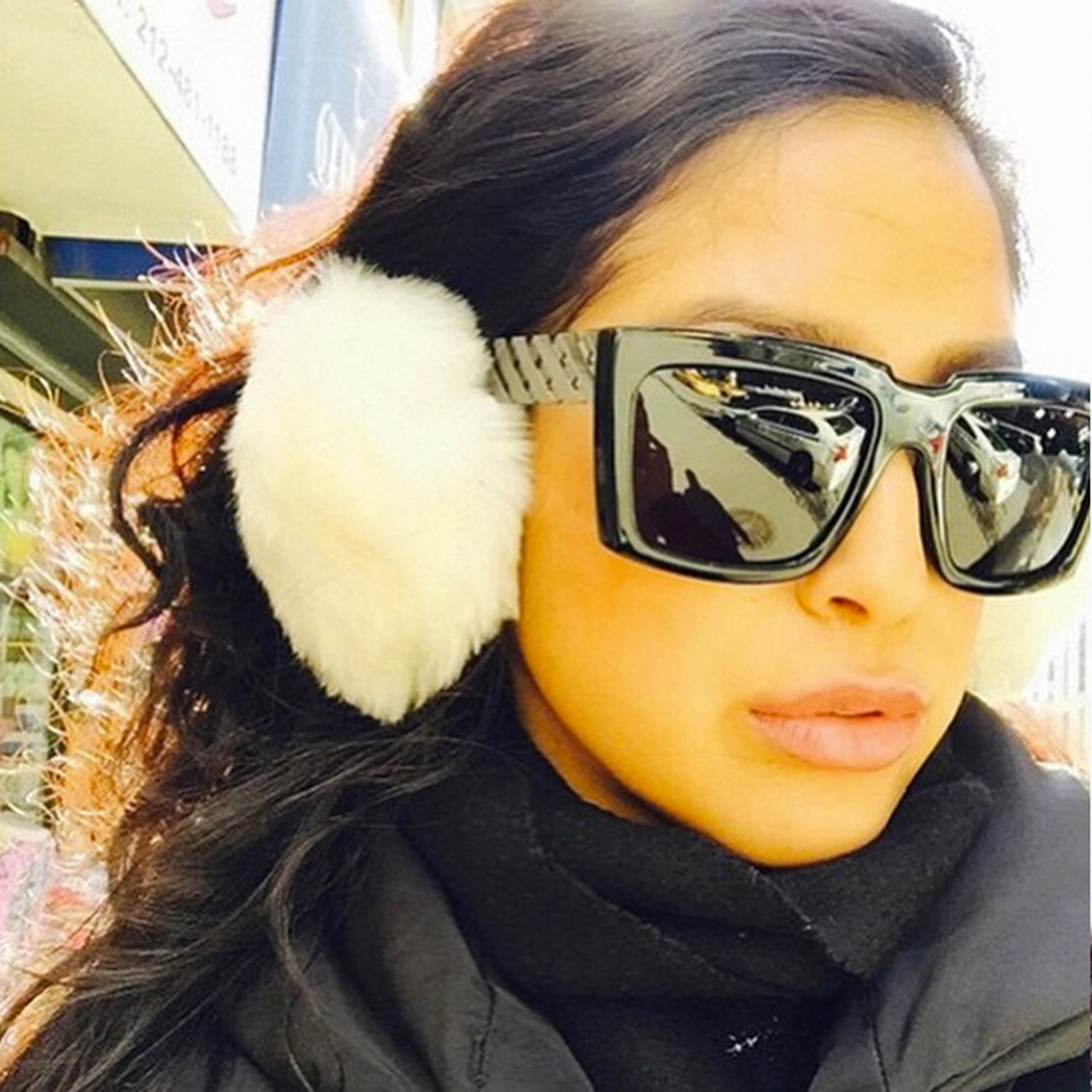 Woman with large sunglasses and white earmuffs in sunlight, wearing a black outfit.