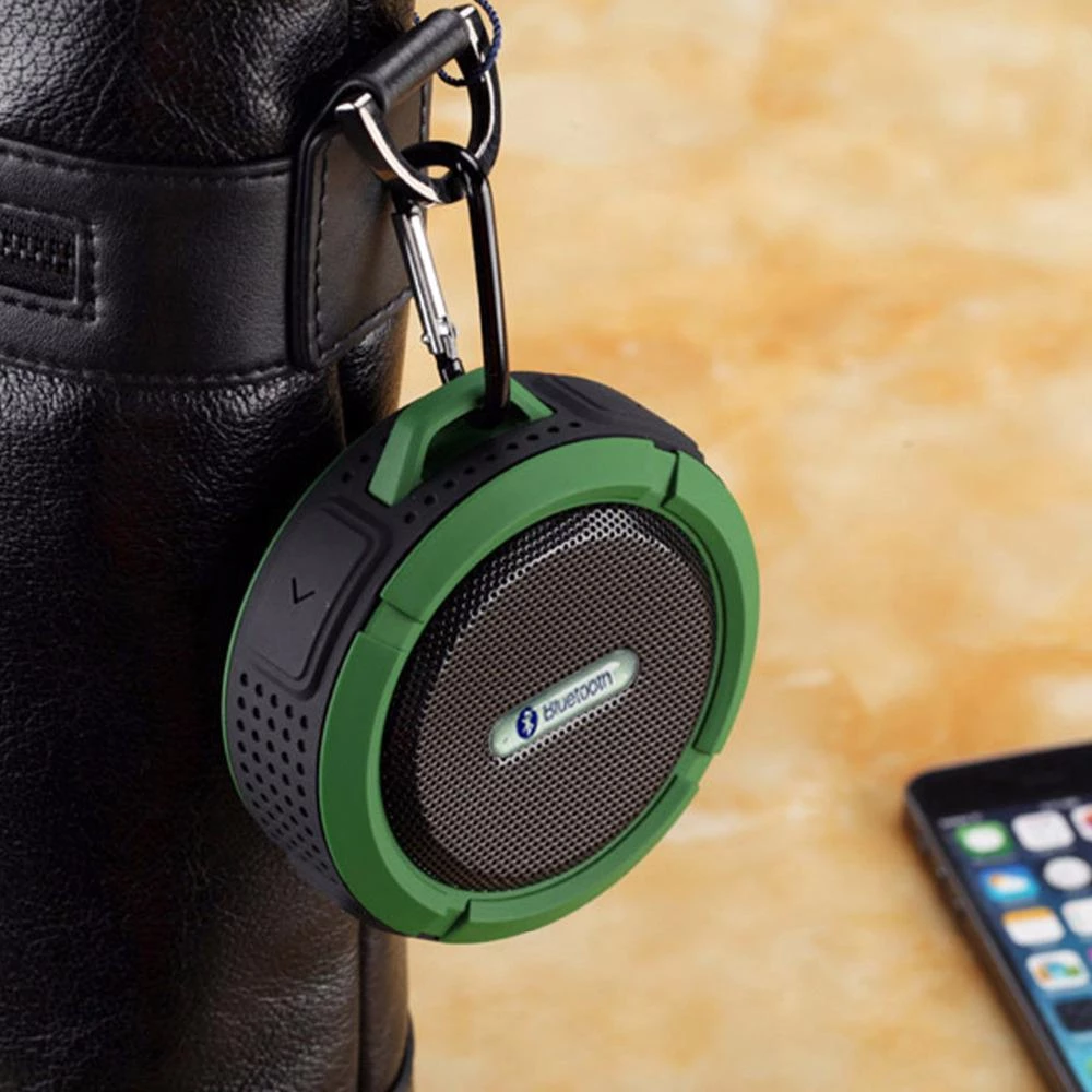 Green and black Bluetooth speaker clipped to a black bag on a tan surface, with a smartphone nearby.