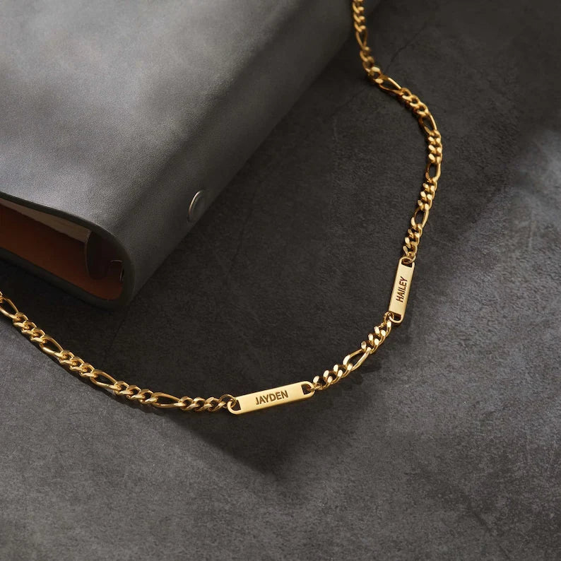 Gold chain with two engraved nameplates on gray surface beside a dark wallet.