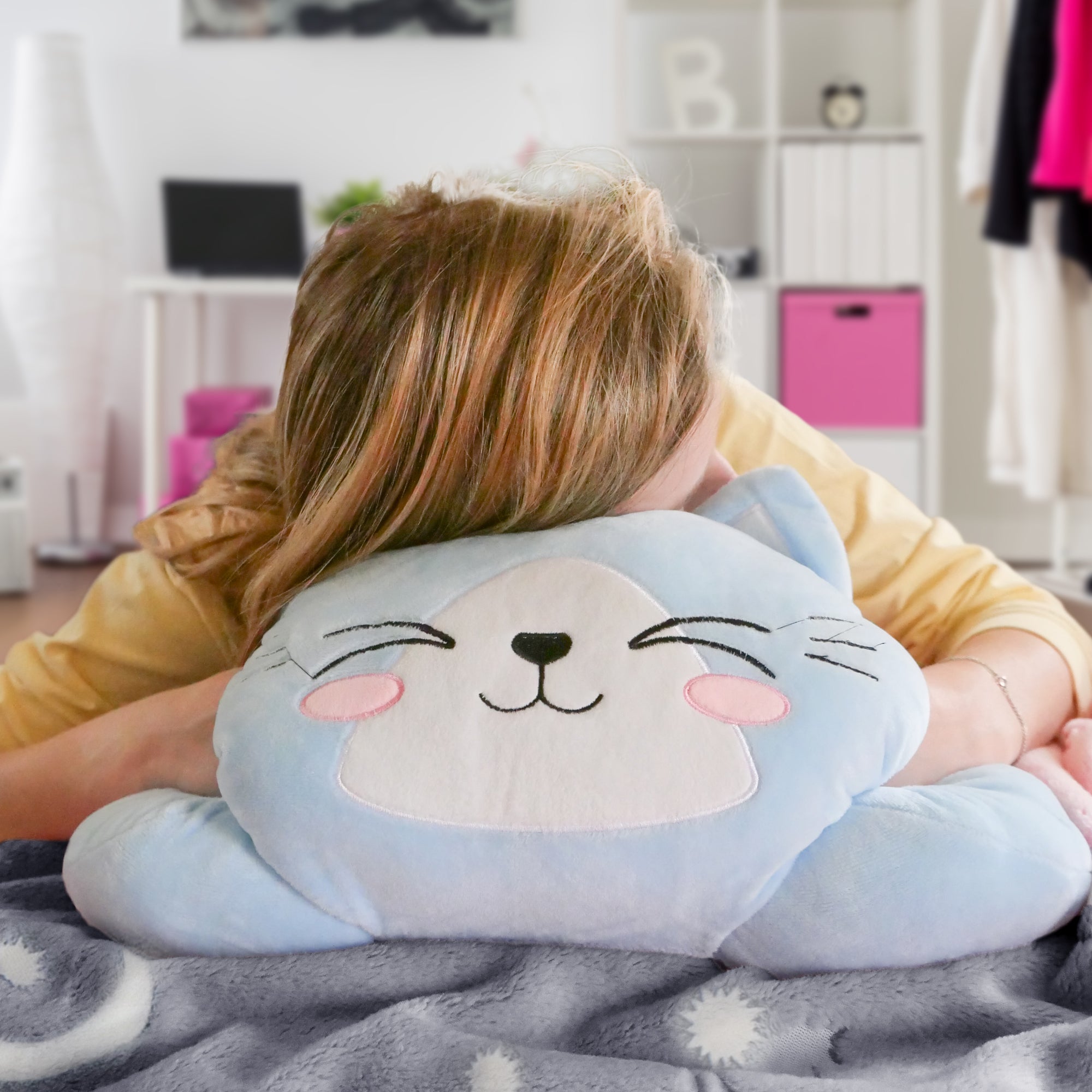 Person hugging a smiling blue cat pillow with pink cheeks, in a cozy room with soft colors.