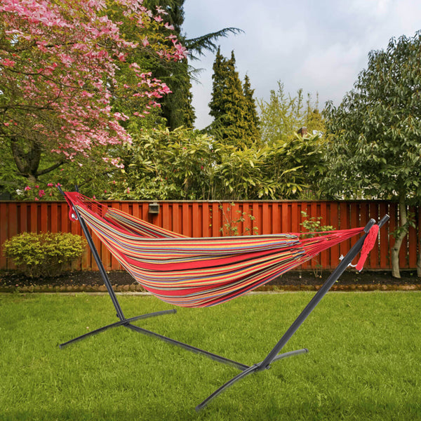 Striped hammock on a metal stand, green lawn, red fence, pink flowering tree, and greenery.