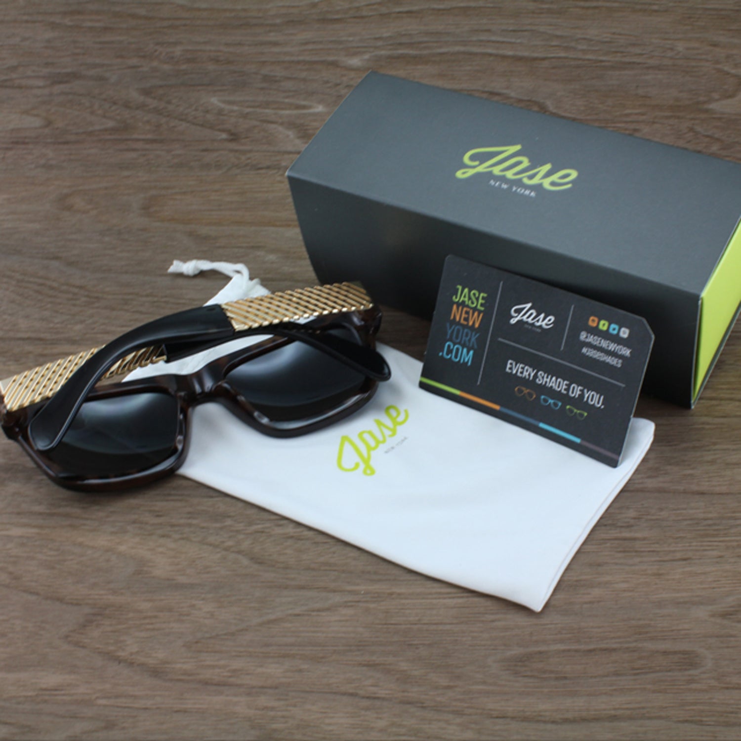 Sunglasses with gold accents, white pouch, gray box, and business card on a wooden surface.