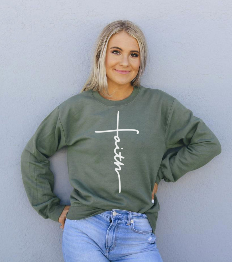 Woman in green "faith" sweatshirt and jeans, stands against a gray wall, smiling, hands on hips.