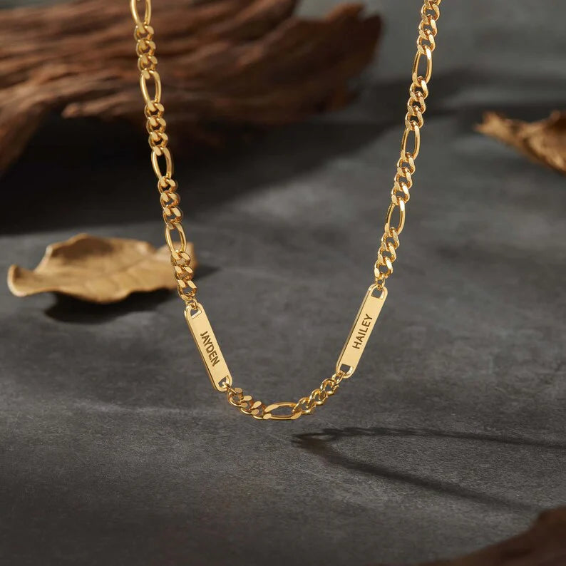 Gold chain necklace with two name plates, "JAYDEN" and "HAILEY," on a dark, textured surface.