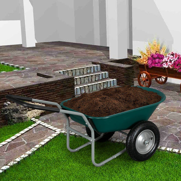 Green wheelbarrow with soil on grassy lawn, near stone steps and flowers in wooden cart.