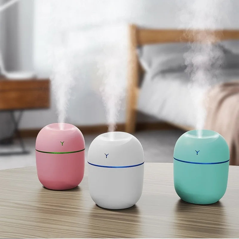 Three colorful humidifiers—pink, white, and teal—emitting mist on a wooden table.