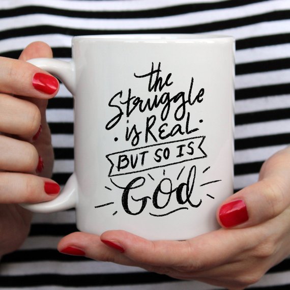 A white mug with black lettering, "The struggle is real, but so is God," held by red-painted nails.