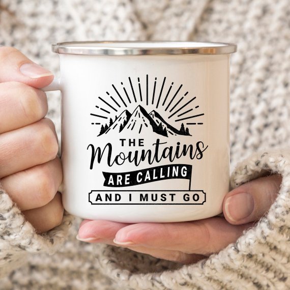 White mug with "The Mountains Are Calling and I Must Go" text, held by hands in a knitted sweater.