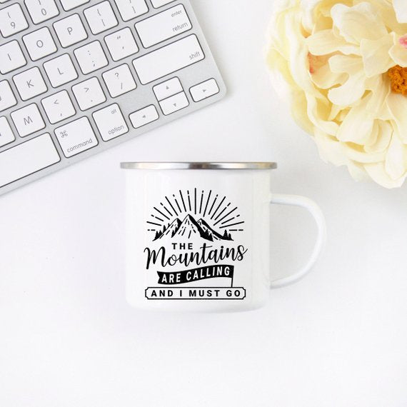White mug with mountain design and text, next to a white keyboard and cream flower.