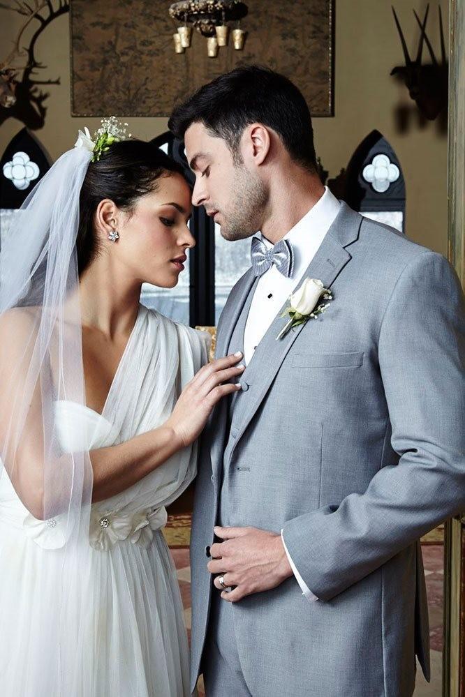 Bride in white gown and veil with groom in a gray suit, sharing an intimate moment indoors.