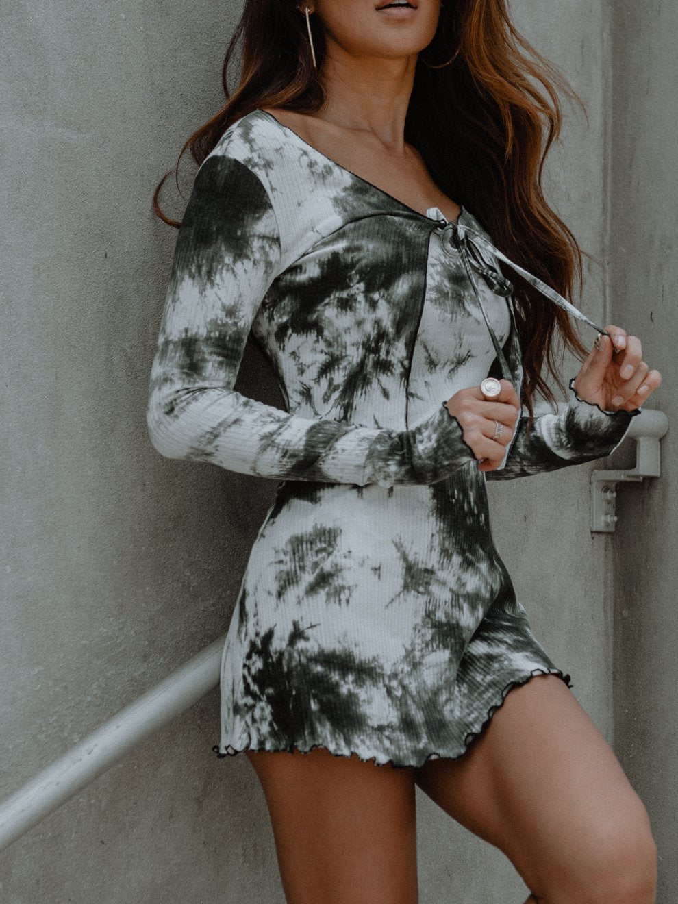 Woman in a black-and-white tie-dye dress with wavy brown hair, leaning against a gray wall.