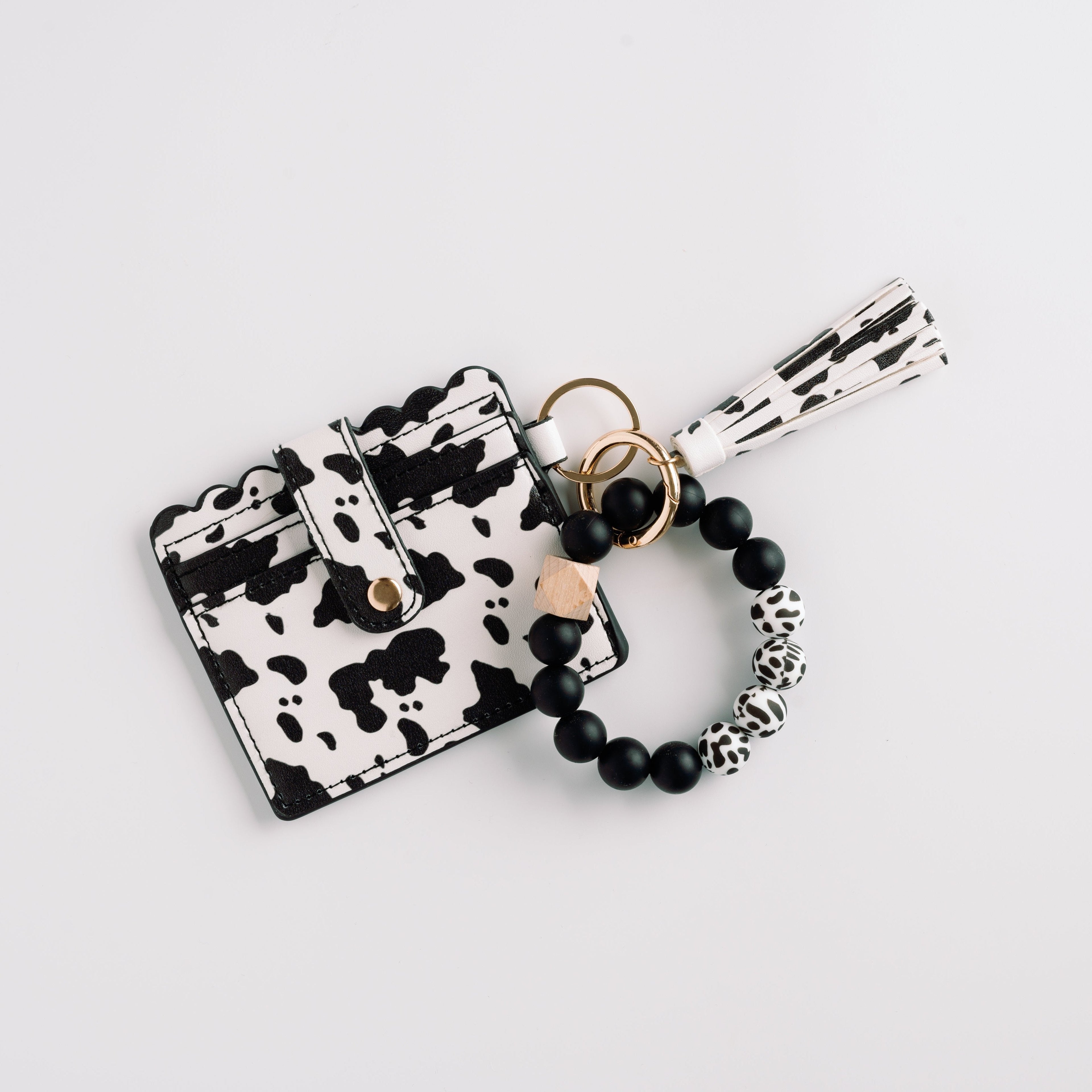Cow-print cardholder with scalloped edges, gold button, and black bead bracelet with tassel.