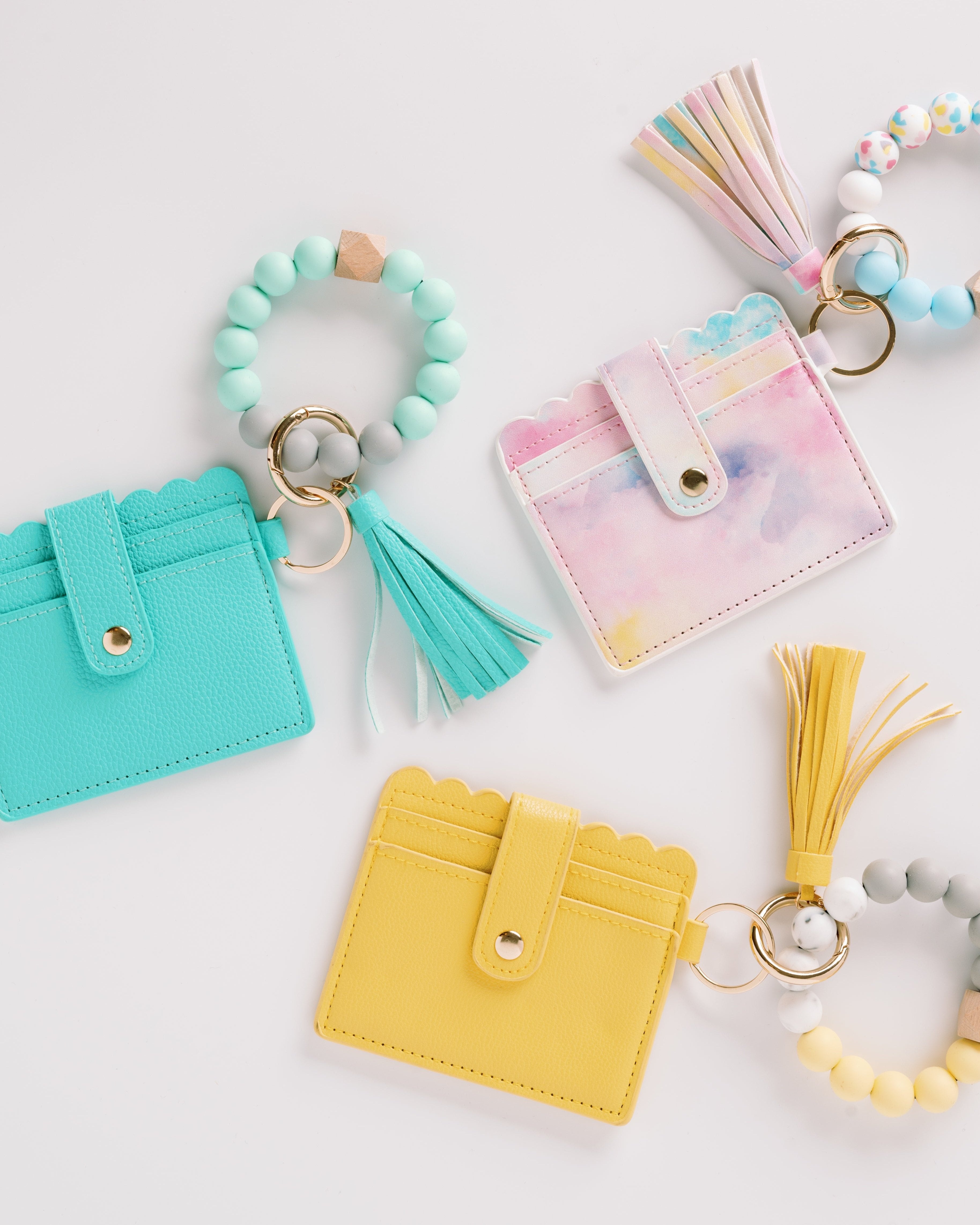 Colorful keychain wallets with tassels and beaded rings in aqua, pastel, and yellow on white.