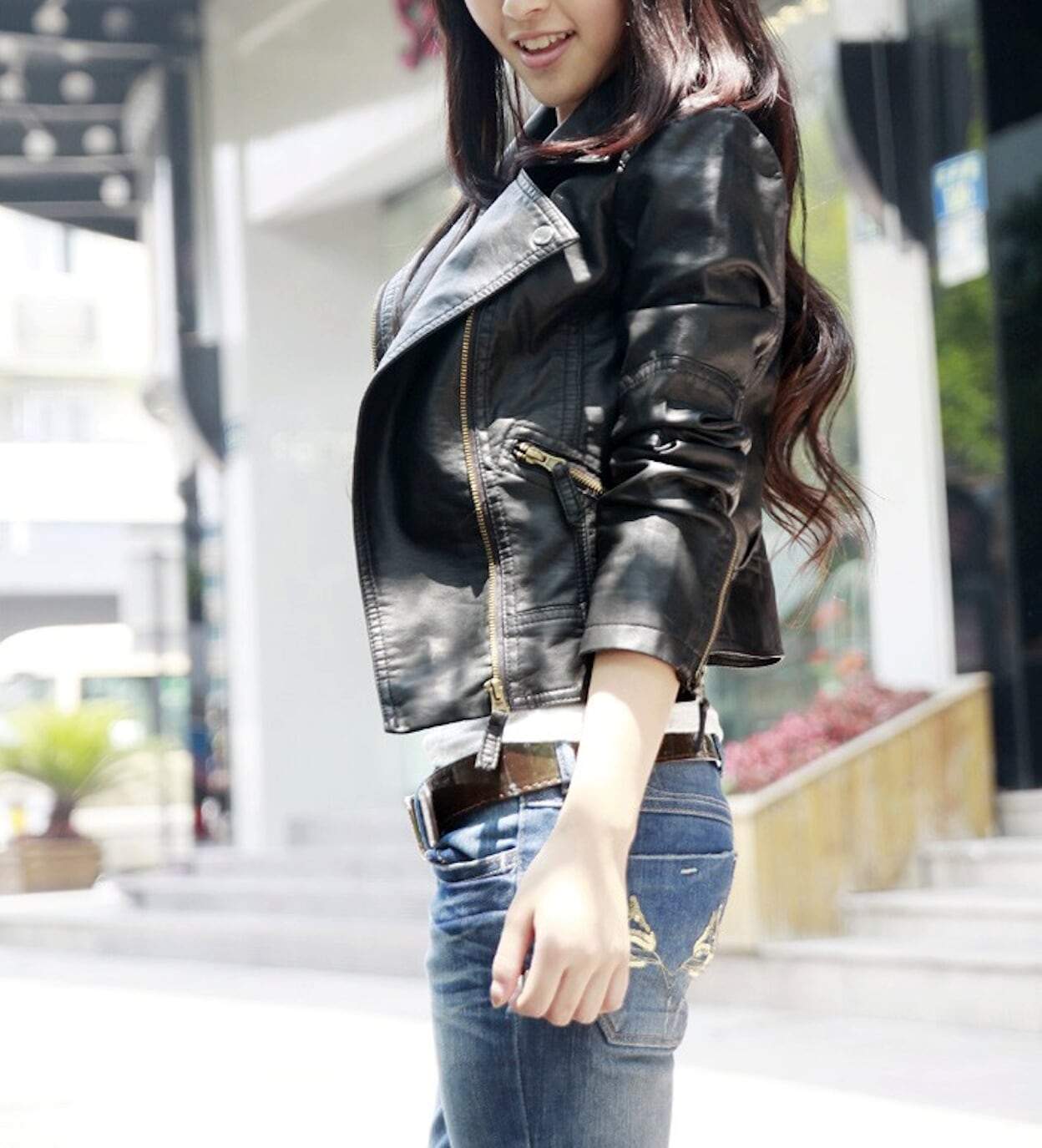 Woman in a black leather jacket and blue jeans walking outdoors, bright sunny day.