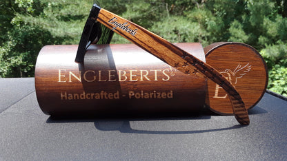 Wooden sunglasses with "Engleberts" on arm rest on a brown engraved case against greenery.