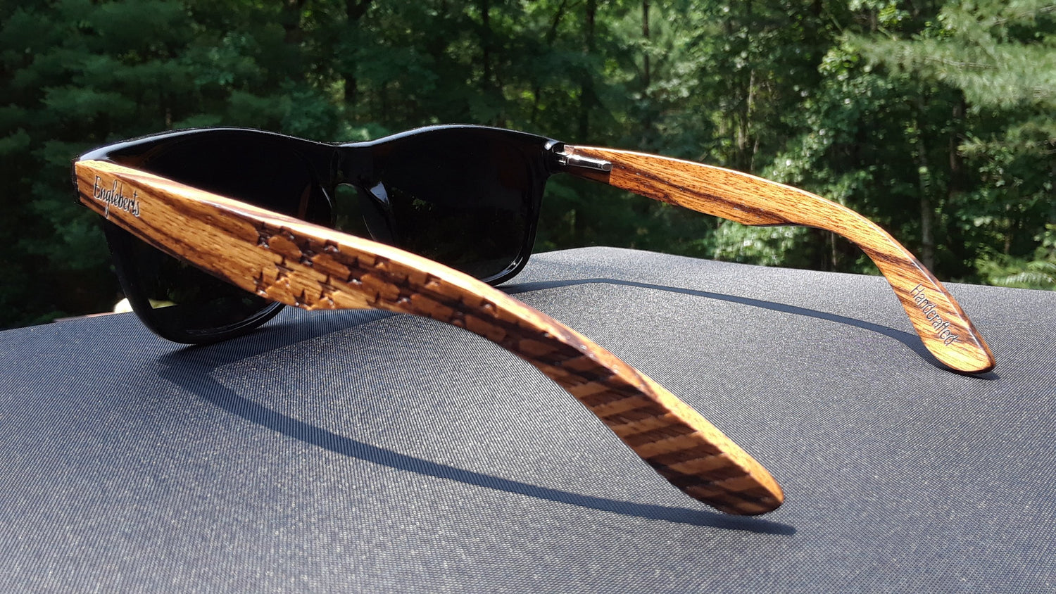 Black sunglasses with striped wooden arms on a dark fabric surface, green forest in background.