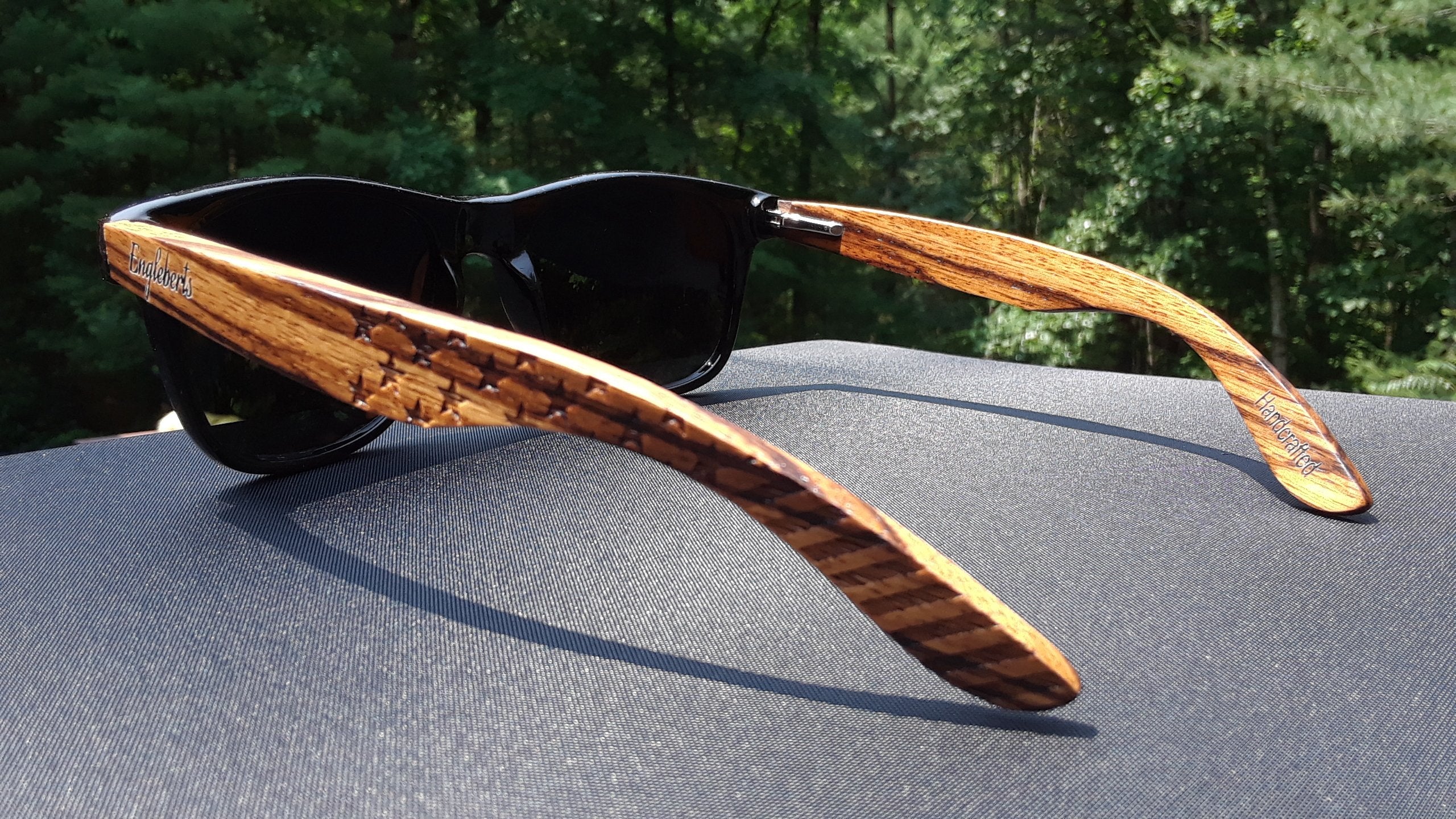 Black sunglasses with striped wooden arms on a dark fabric surface, green forest in background.