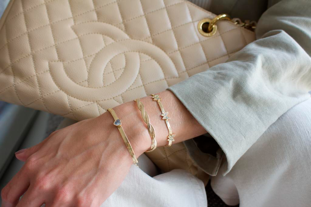 A hand with gold bracelets rests on a beige quilted bag with a gold chain, wearing light green sleeves.