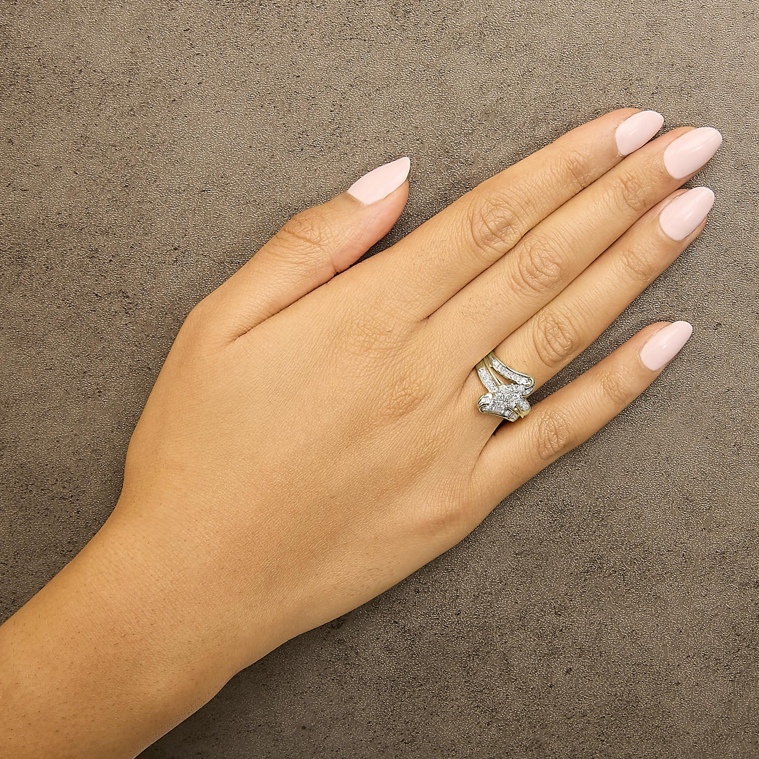 Hand with light pink nails on a textured surface, wearing a gold ring with a large diamond flower.