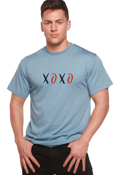 Man wearing a light blue T-shirt with "XOXO" in black and red letters, hands in pockets.