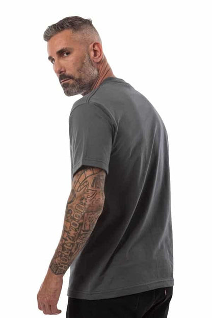 Man with short hair wearing a gray shirt and displaying a tattooed arm, looking over his shoulder.