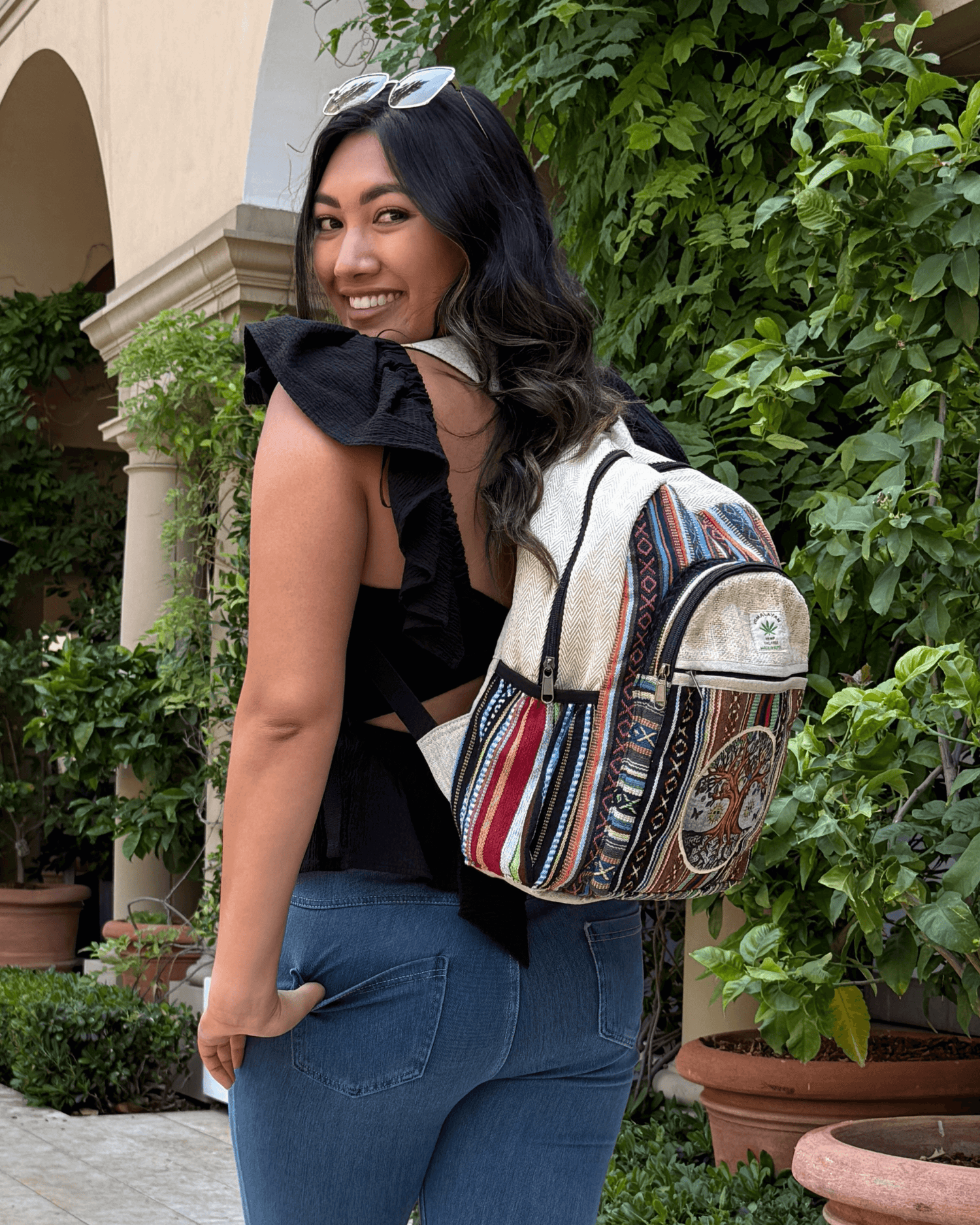 Smiling woman in black top with colorful backpack, standing by leafy plants and archway.