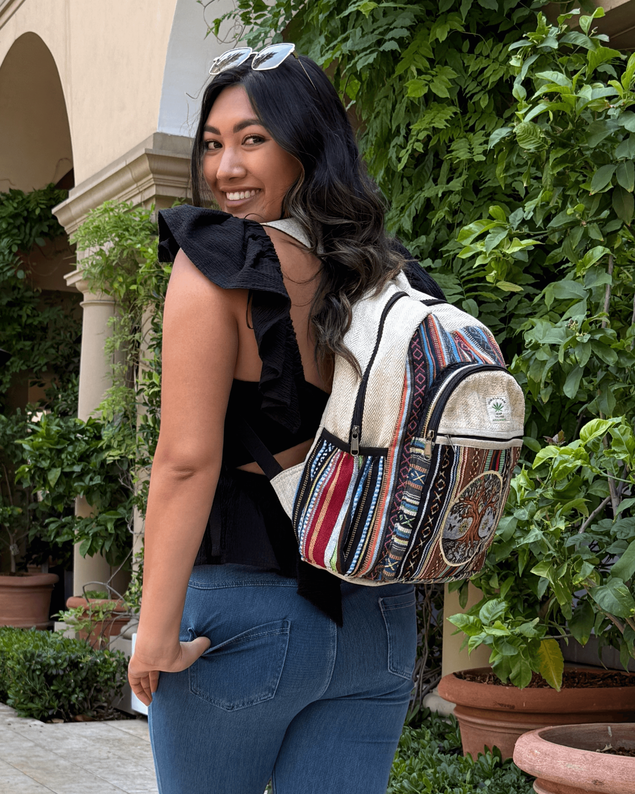 Smiling woman in black top with colorful backpack, standing by leafy plants and archway.