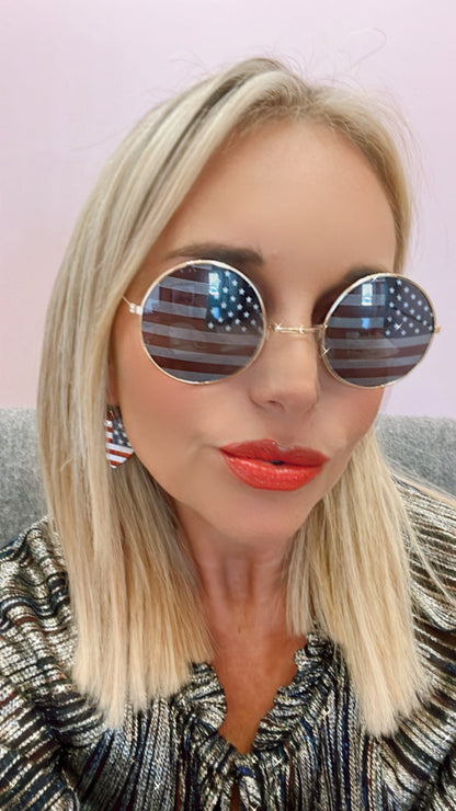 A woman wears sunglasses with American flag reflections, red lipstick, and a striped blouse.