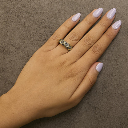 Hand with lilac nails wearing a gold and black patterned ring on a textured gray surface.
