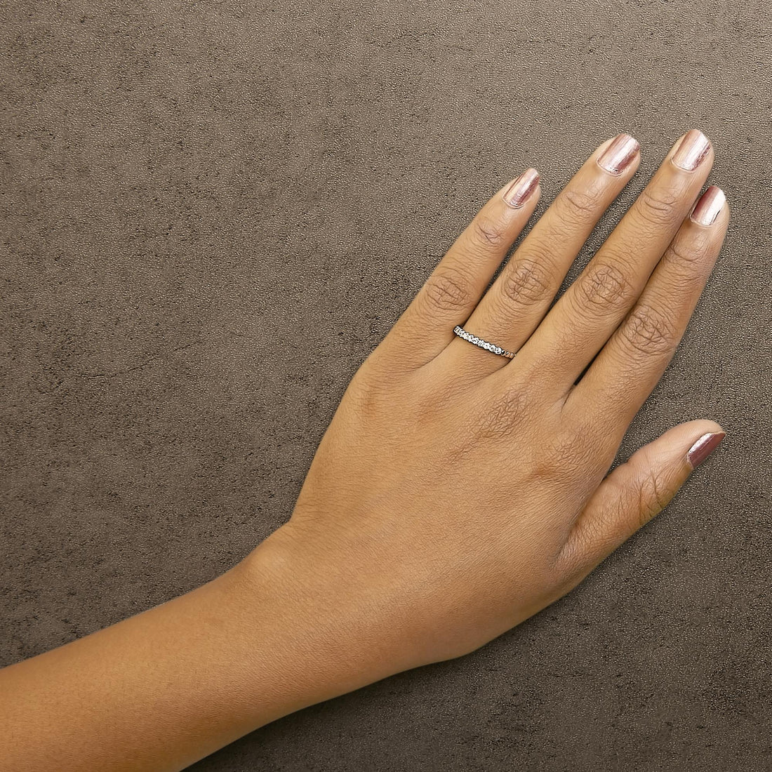 A hand with metallic pink nails wearing a diamond band ring on a textured brown background.
