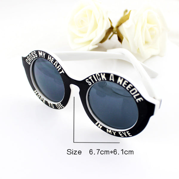 Round black sunglasses with white text, white frame, and dark lenses; white roses in background.