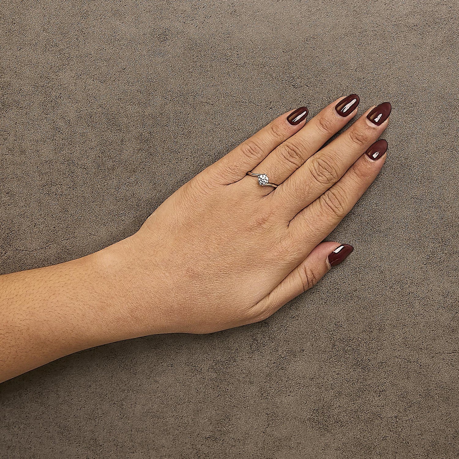 Hand with dark burgundy nails wearing a silver ring with a diamond on a textured brown surface.