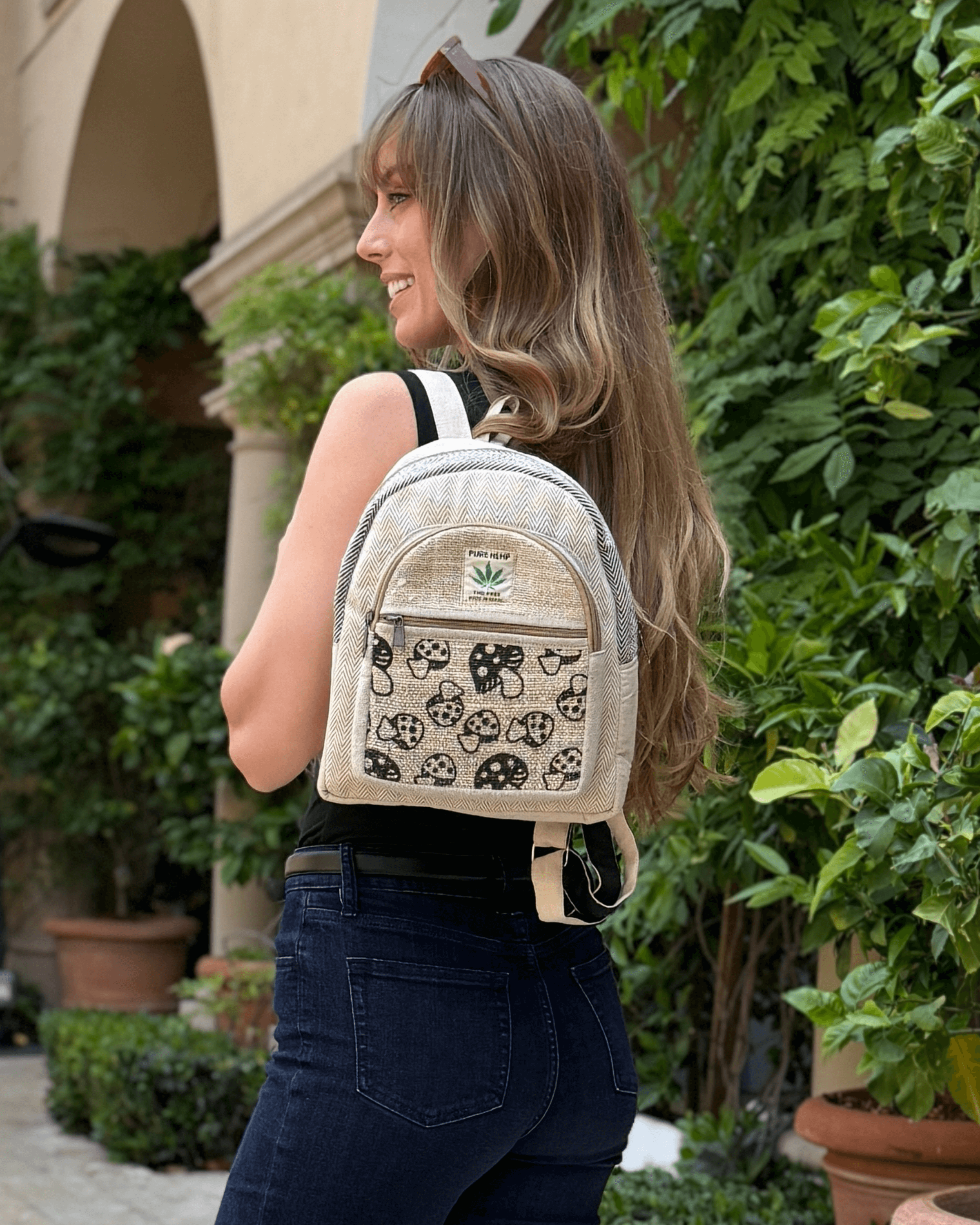Woman with long hair wearing a black top, jeans, and a patterned beige backpack, standing outside.