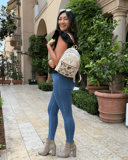 Woman with a beige backpack, wearing blue jeans and a black top, stands smiling on a tiled path.